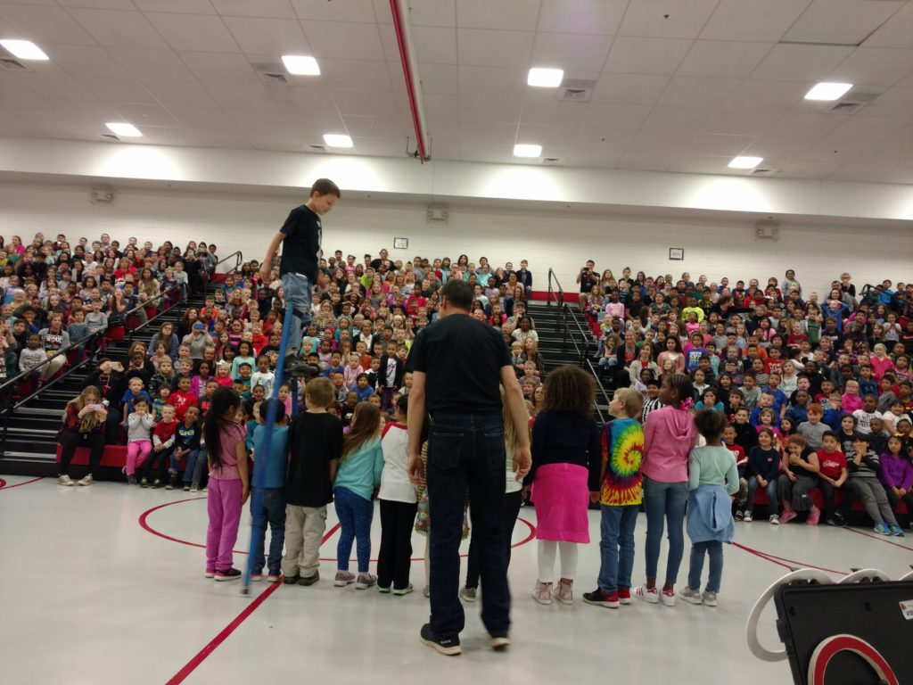 Nathan on stilts in front of a gym packed with kids